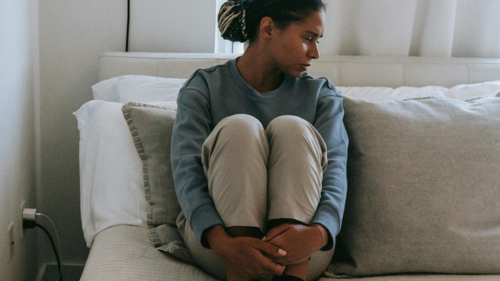 Woman sitting on a bed holding her knees with a concerned expression, looking to the side.